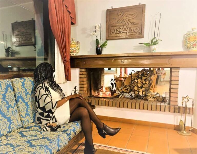 A woman sitting next to a wood fireplace. It represents a low-budget winter destination in Italy