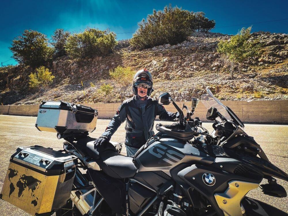 A motorcyclist standing behind his motorcycle with the backdrop of a grassy hill. This is to show accessories that can be used as gift ideas for a motorcycle lover