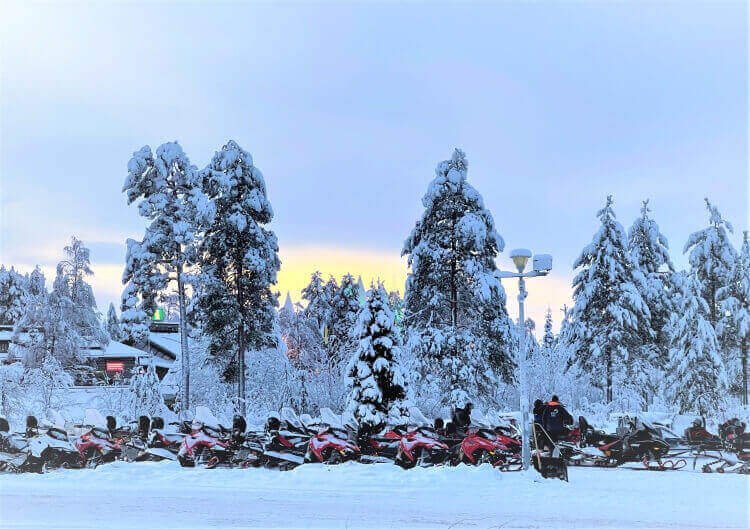 A winter landscape with tall snow-covered trees and snowmobiles, and a sunset behind in Rovaniemi Lapland Complete Guide