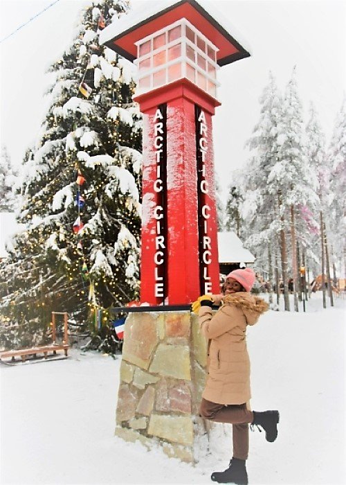 A woman standing next to a pillar with the words "Arctic Circle" written vertically, in Rovaniemi Lapland