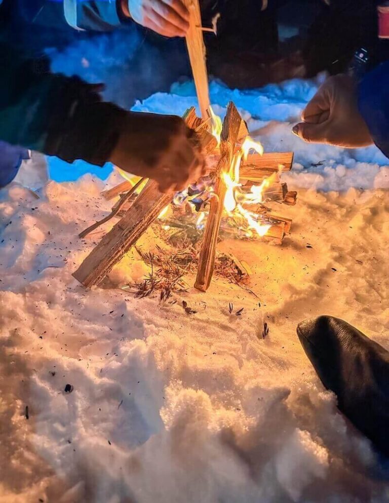 Several hands adding wood to start a fire on a snow-covered ground in Rovaniemi Lapland