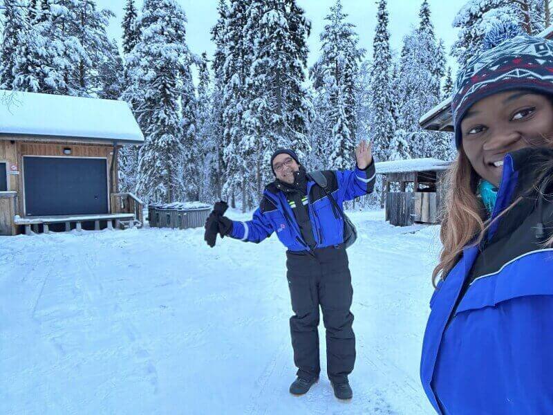 A couple dressed in winter overalls in a winter landscape with tall pine trees covered in snow in Rovaniemi Lapland