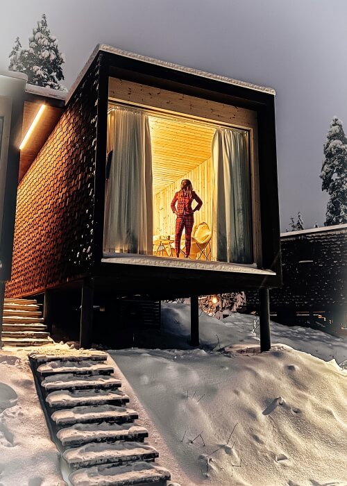 A woman standing in the glass window of a wooden cabin in a winter landscape in Rovaniemi Lapland